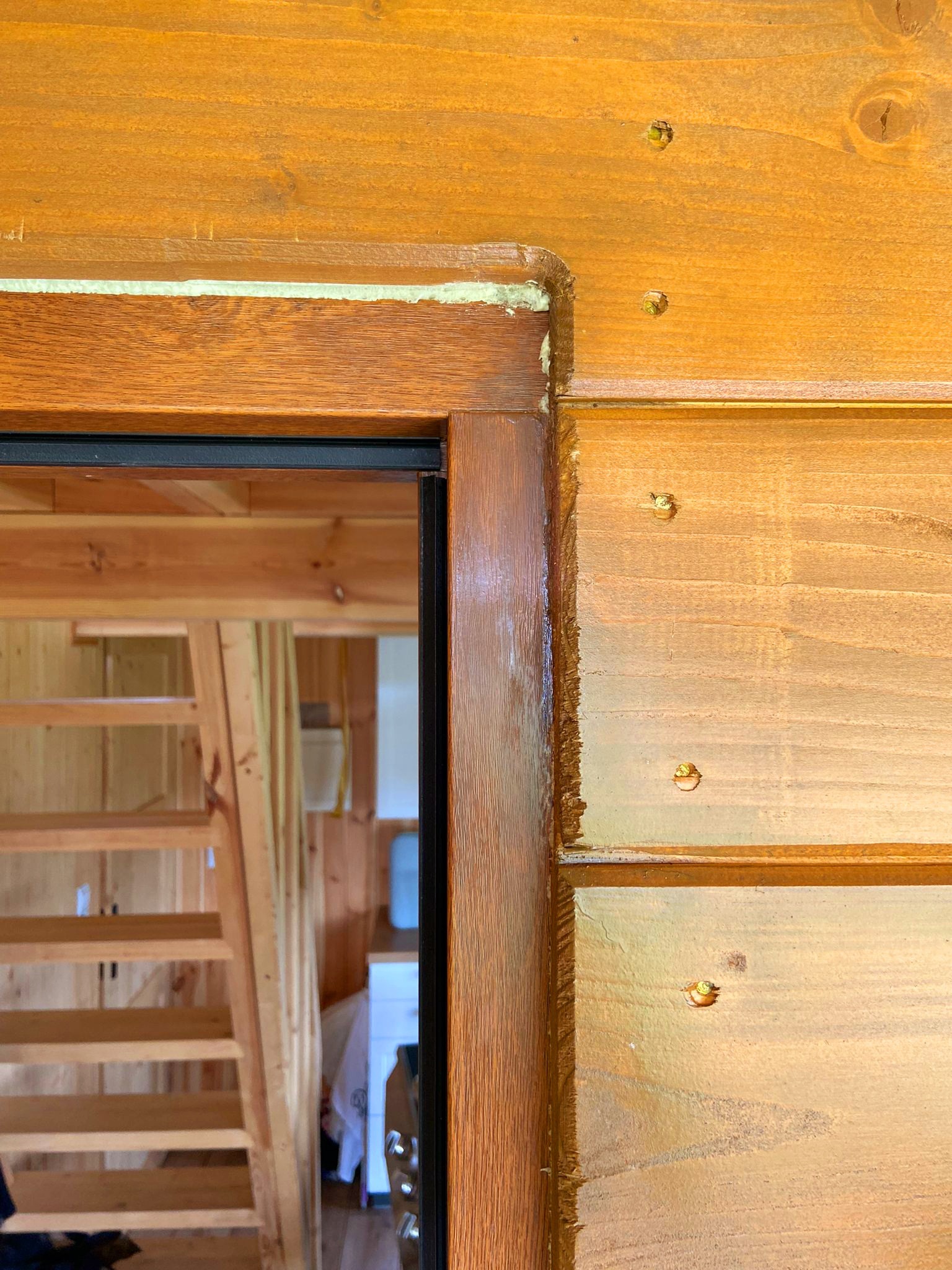 Fragment drewnianej ościeżnicy i ściany z widocznymi śladami świeżo nałożonego uszczelniacza, z widocznym w tle fragmentem drewnianych schodów.