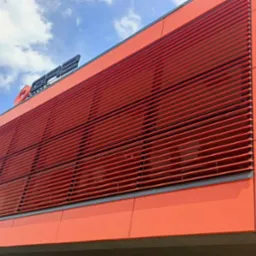 Pomarańczowy budynek z poziomymi, czerwonymi żaluzjami zewnętrznymi i logo firmy na dachu, widziany z dołu pod kątem.
