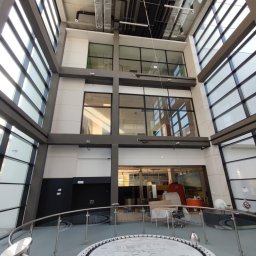 IndustriaAlp Kamil Szewc - Wnętrze nowoczesnego budynku z dużymi szklanymi oknami i metalowymi rurami wentylacyjnymi pod sufitem, widoczne elementy wyposażenia i instalacje.