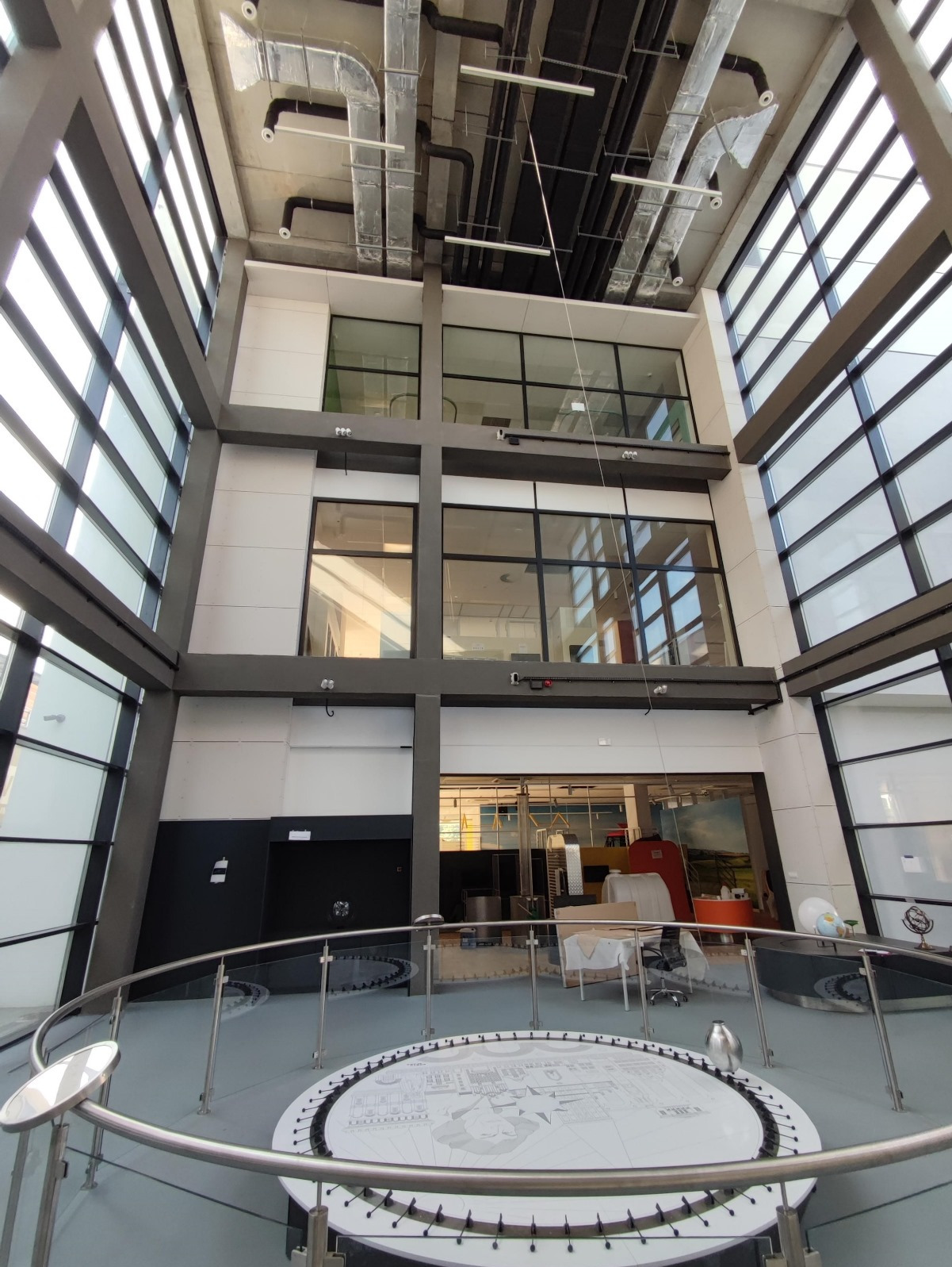 Wnętrze nowoczesnego budynku z dużymi szklanymi oknami i metalowymi rurami wentylacyjnymi pod sufitem, widoczne elementy wyposażenia i instalacje.