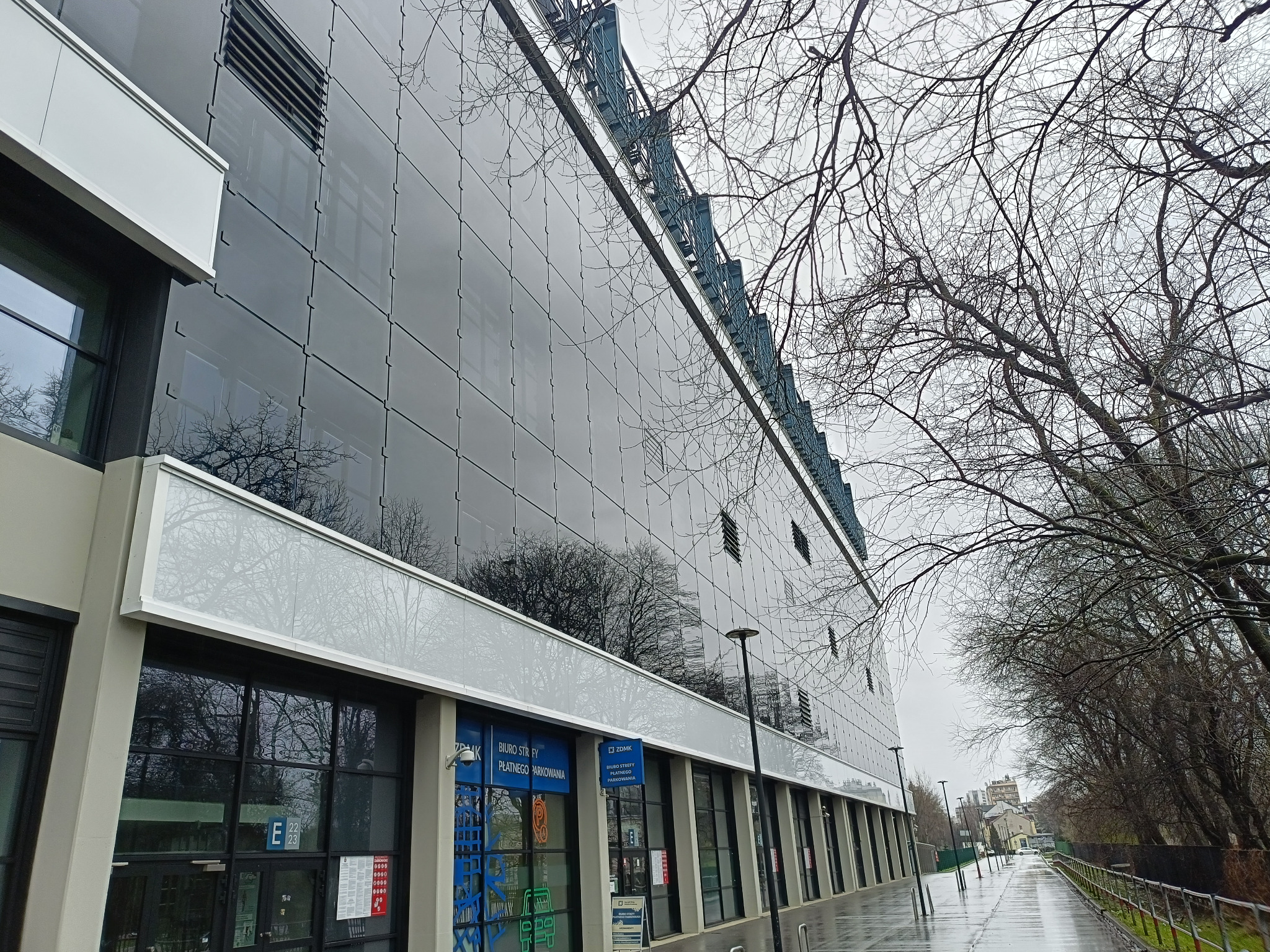 Szklana elewacja budynku biurowego odbijająca nagie drzewa w pochmurny dzień po deszczu, widok z poziomu chodnika.