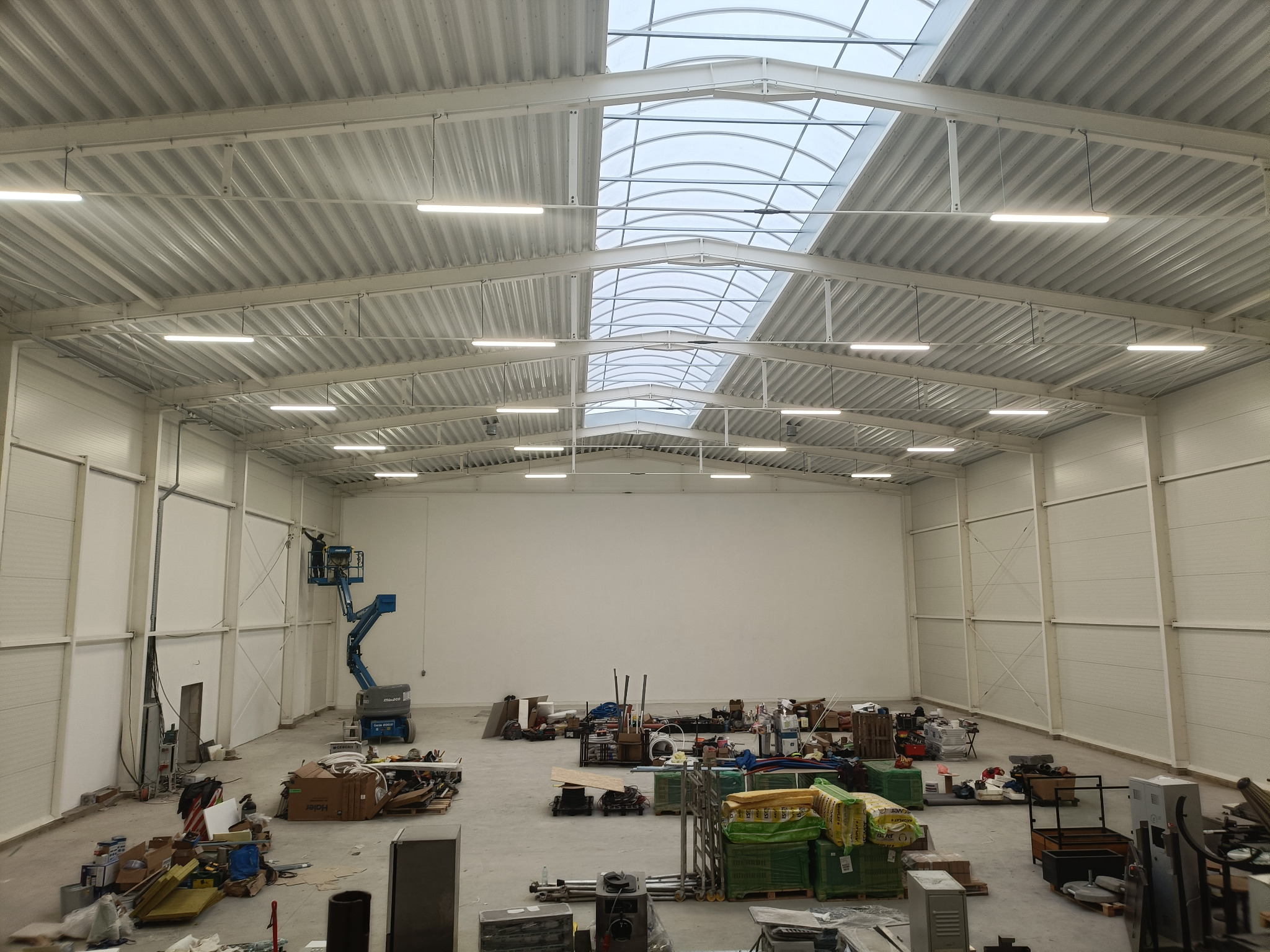 Wnętrze hali przemysłowej w trakcie budowy, widoczna konstrukcja dachu z oknami dachowymi, oświetlenie LED, niebieski podnośnik koszowy oraz materiały budowlane na podłodze.