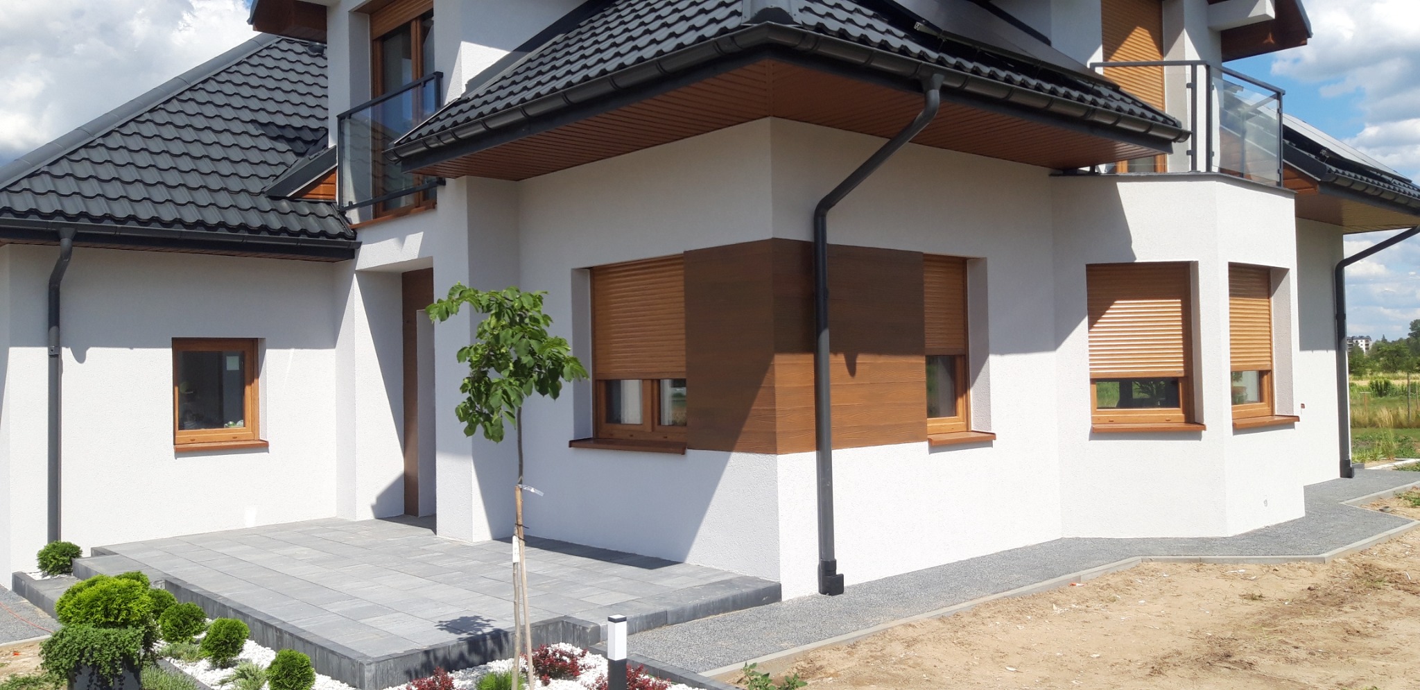 Nowoczesny dom jednorodzinny z białą elewacją, drewnianymi okiennicami w kolorze jasnego brązu, czarną dachówką i systemem rynnowym, oraz szarą kostką brukową na podjeździe i wokół budynku.