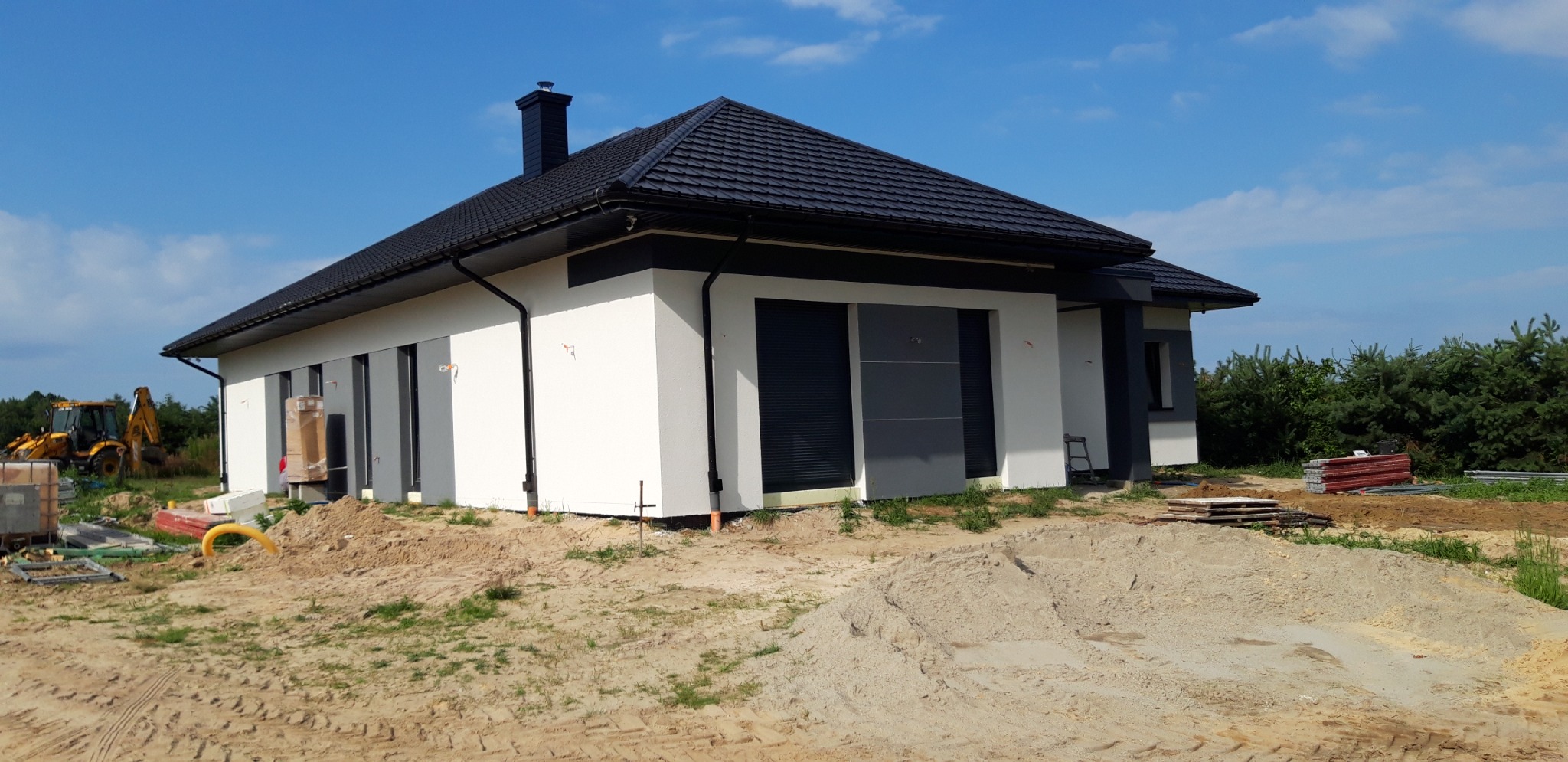 Nowoczesny dom jednorodzinny z białą elewacją i czarnymi akcentami, czarną dachówką, widoczny na etapie prac wykończeniowych, z żółtą koparką w tle i stertą piachu na pierwszym planie.