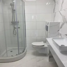 Nowoczesna łazienka z kabiną prysznicową, podwieszaną toaletą i blatem z umywalką, wykończona białymi płytkami imitującymi marmur.