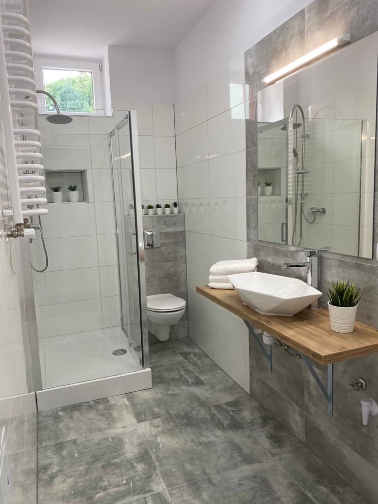Nowoczesna łazienka z kabiną prysznicową, umywalką nablatową o geometrycznym kształcie na drewnianym blacie i szarymi płytkami imitującymi kamień na podłodze i częściowo na ścianach.