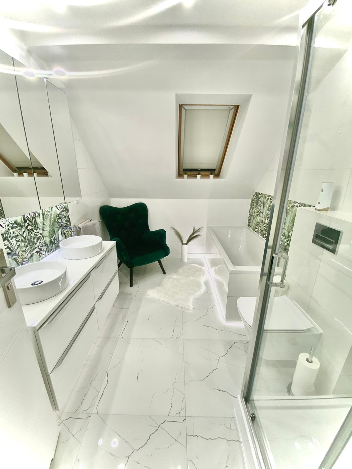 Jasna łazienka na poddaszu z marmurową posadzką, dwoma umywalkami na białej szafce, wanną z podświetleniem LED i zielonym fotelem.