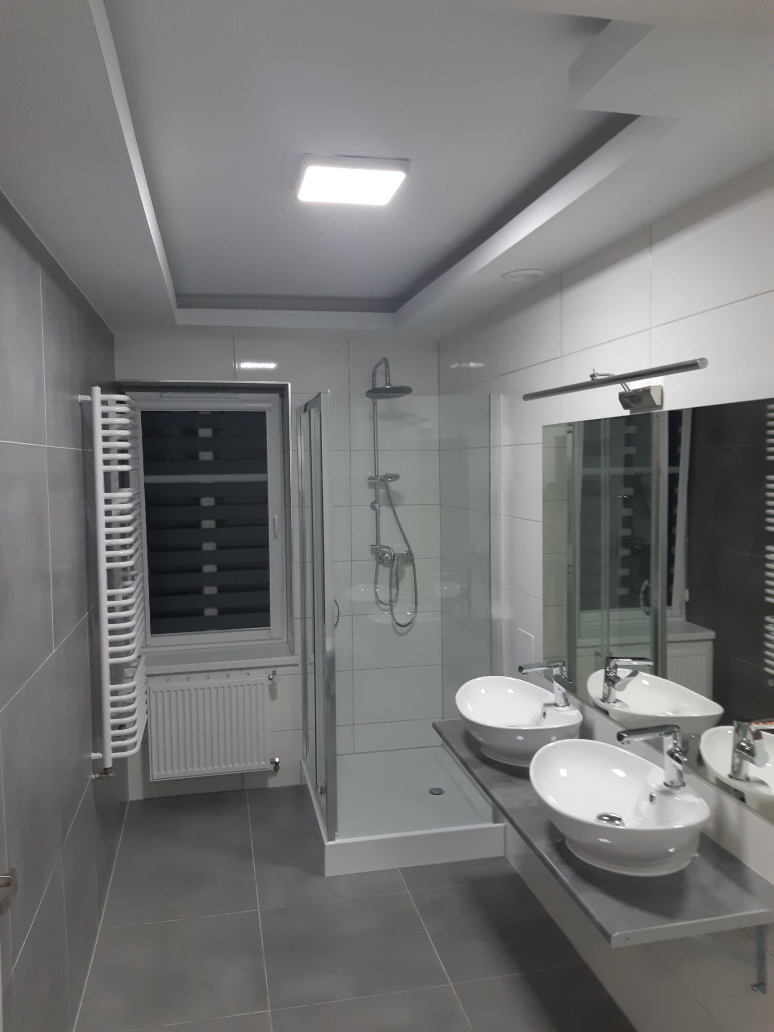Wykończona łazienka w odcieniach szarości i bieli z dwoma umywalkami nablatowymi, kabiną prysznicową typu walk-in, grzejnikiem drabinkowym i roletami w oknie.