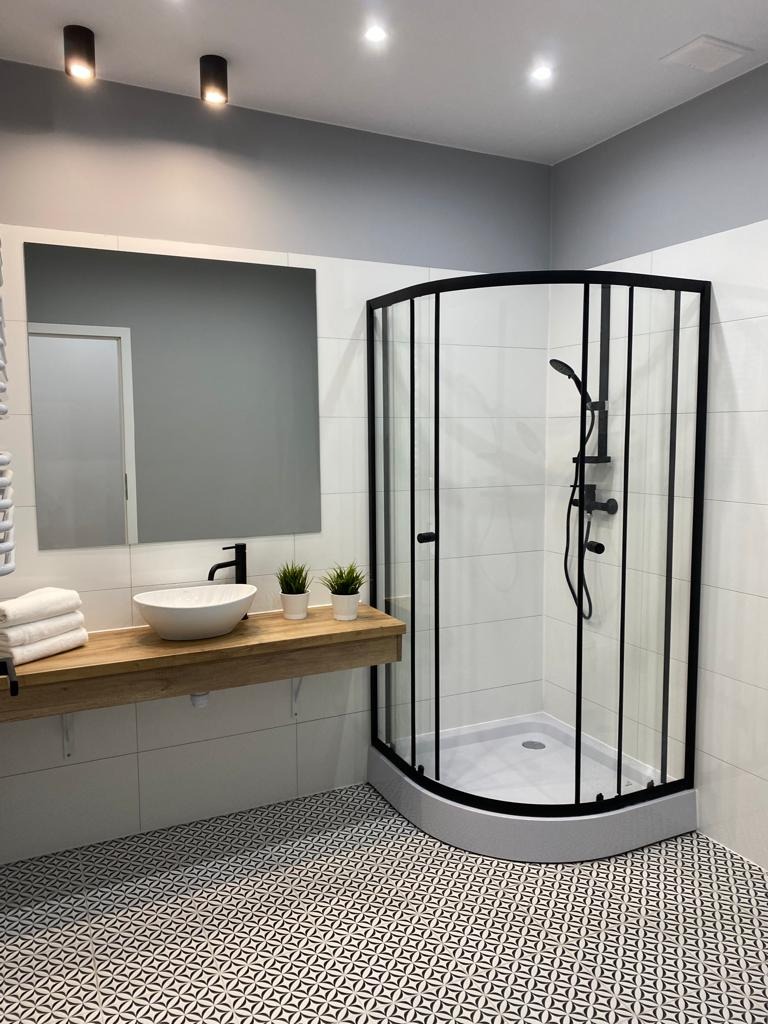 Nowoczesna łazienka z kabiną prysznicową w czarnej ramie, umywalką nablatową na drewnianym blacie, wzorzystą podłogą i białymi płytkami.