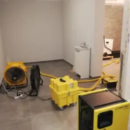 Pokój po zalaniu z widocznymi śladami wilgoci na ścianach, osuszany za pomocą żółtych osuszaczy powietrza marki Trotec i wentylatorów, z widocznymi żółtymi wężami odprowadzającymi wilgoć.