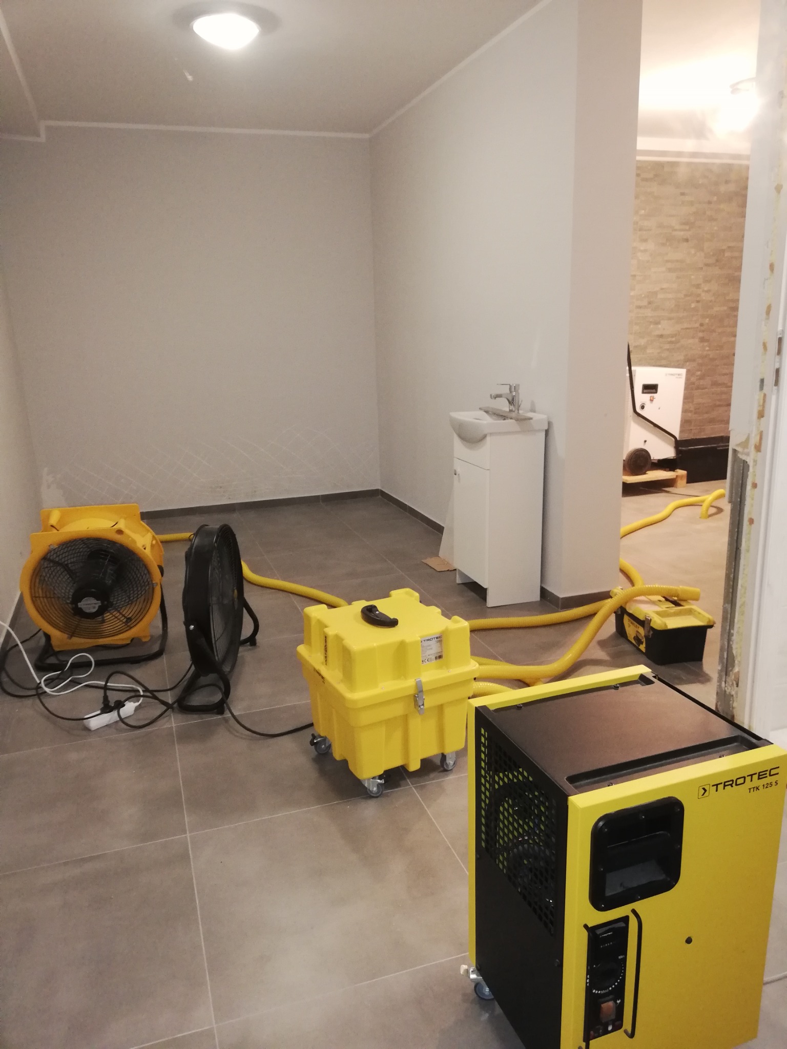Pokój po zalaniu z widocznymi śladami wilgoci na ścianach, osuszany za pomocą żółtych osuszaczy powietrza marki Trotec i wentylatorów, z widocznymi żółtymi wężami odprowadzającymi wilgoć.