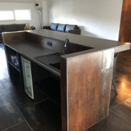 Wnętrze z wyspą kuchenną wykończoną panelami imitującymi rdzę, zintegrowaną z barkiem i małą lodówką na wino, na tle salonu z szarą sofą.