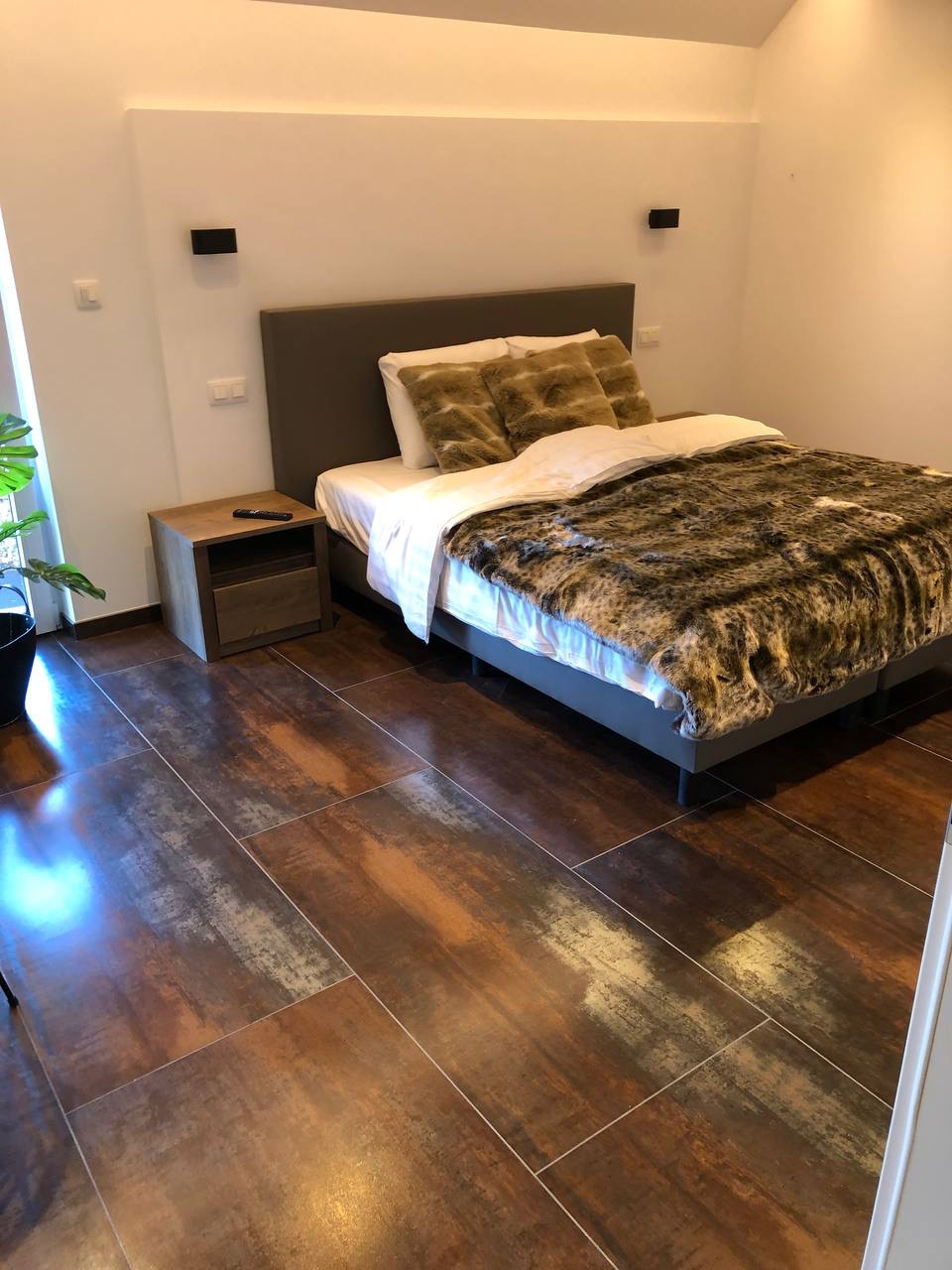 Sypialnia z łóżkiem z szarym zagłówkiem, wzorzystą narzutą i poduszkami w odcieniach brązu, drewniany stolik nocny z pilotem, ciemne płytki podłogowe imitujące drewno.