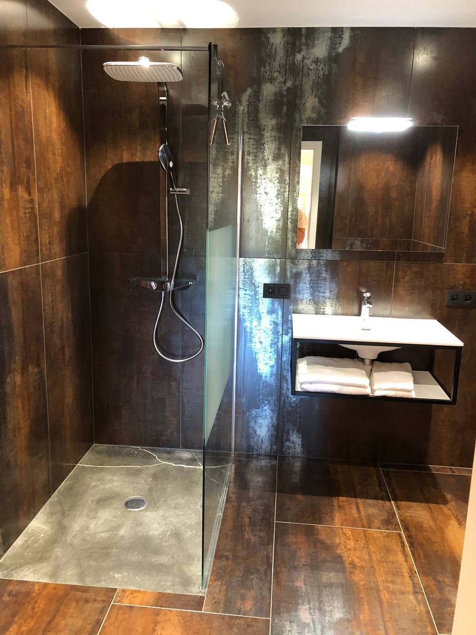 Nowoczesna łazienka z kabiną prysznicową typu walk-in, wykończona płytkami imitującymi rdzę, z betonową posadzką w strefie prysznica i minimalistyczną umywalką podświetloną lampą.