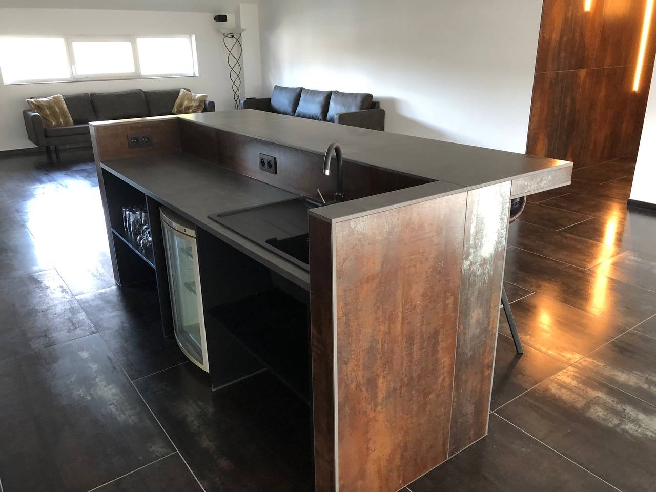 Wnętrze z wyspą kuchenną wykończoną panelami imitującymi rdzę, zintegrowaną z barkiem i małą lodówką na wino, na tle salonu z szarą sofą.