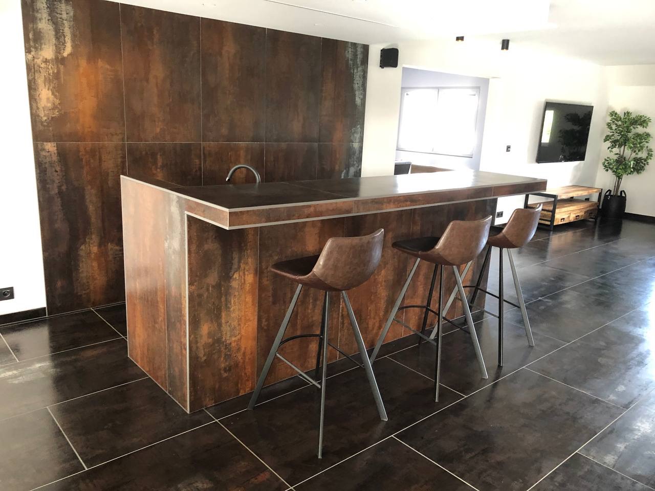 Rustykalny bar z płytek imitujących rdzę, z trzema skórzanymi hokerami na tle salonu z telewizorem i rośliną doniczkową.