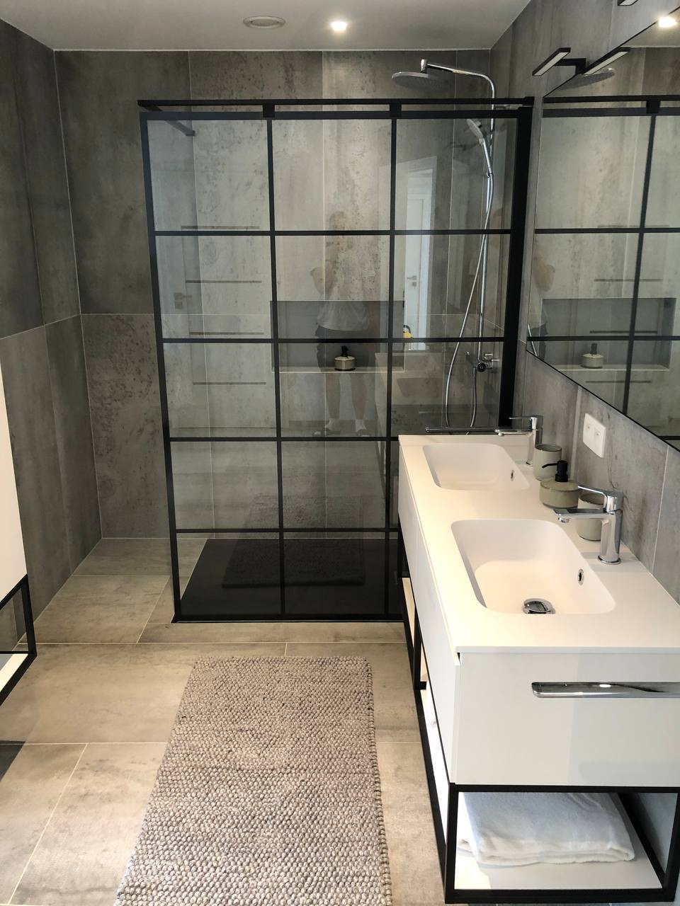 Nowoczesna łazienka z kabiną prysznicową typu walk-in z czarnymi ramami, białą podwójną umywalką i szarą płytką na ścianach oraz podłodze, uzupełniona o dywanik.