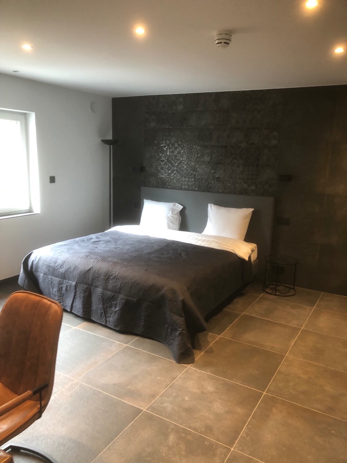 Nowoczesna sypialnia z ciemną, teksturowaną ścianą za łóżkiem, szarą pościelą i płytkami podłogowymi w odcieniach szarości i brązu. Widoczne oświetlenie punktowe w suficie i fragment brązowego...