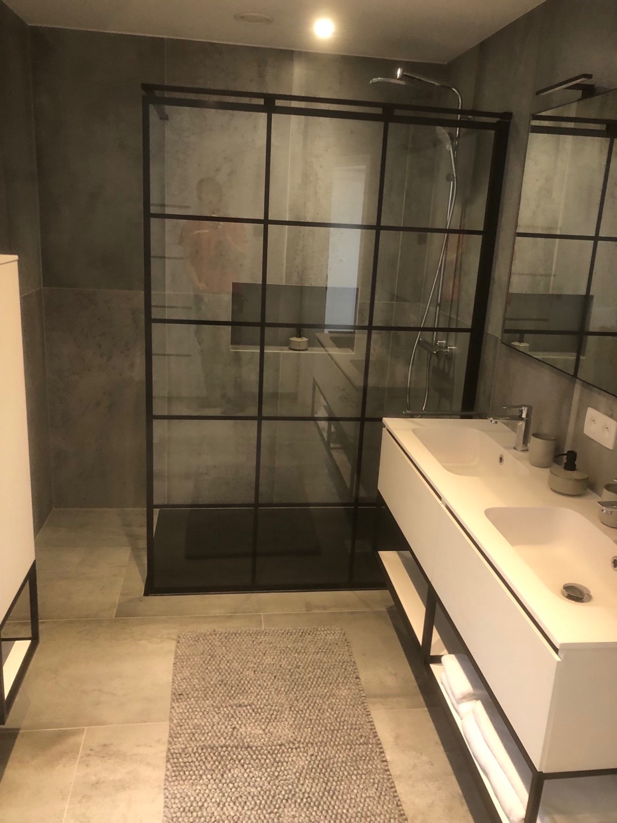 Nowoczesna łazienka z kabiną prysznicową typu walk-in z czarnymi profilami, podwójną białą umywalką na szafce z otwartymi półkami i szarymi płytkami na ścianach.