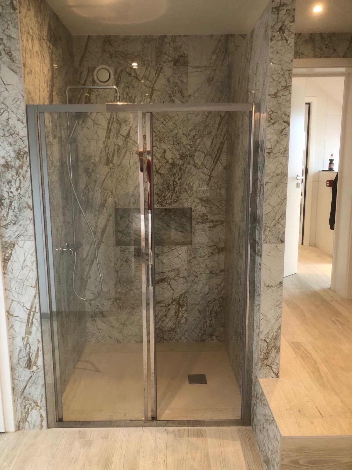 Kabina prysznicowa z szarymi marmurkowymi płytkami, deszczownica i półka w ścianie, widok na fragment drewnianej podłogi i otwarte drzwi do innego pomieszczenia.