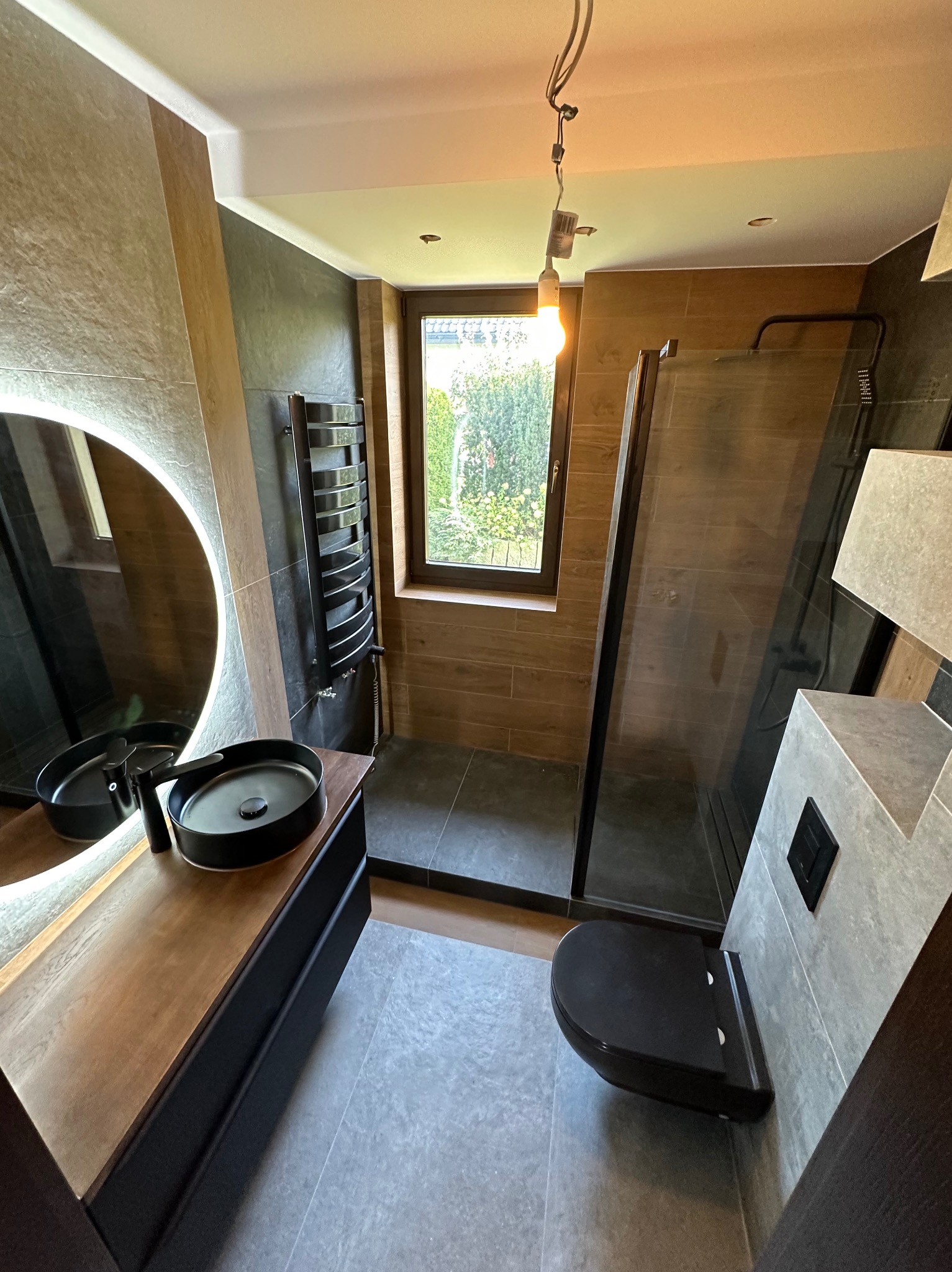 Nowoczesna łazienka z czarną armaturą: okrągłe lustro z podświetleniem, umywalka nablatowa, grzejnik drabinkowy i kabina prysznicowa typu walk-in z widokiem na zieleń za oknem.