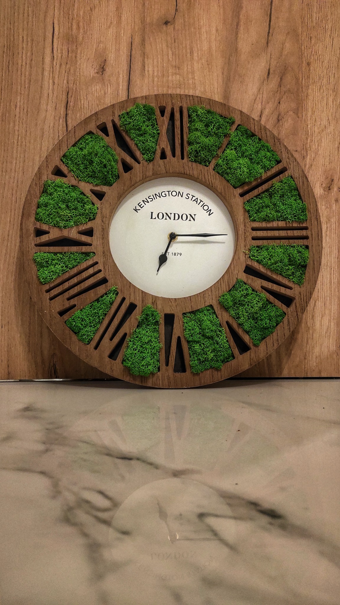 Okrągły zegar ścienny z drewnianą ramą i mchem w miejscu cyfr rzymskich, tarcza z napisem Kensington Station London, odbicie zegara na błyszczącej powierzchni