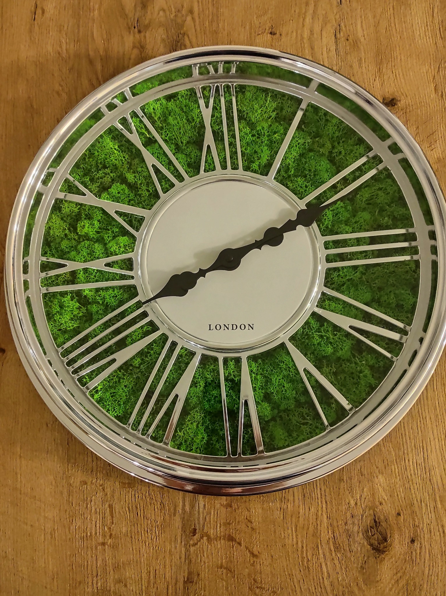 Okrągły zegar ścienny z srebrną ramą i wypełnieniem z zielonego mchu, wskazujący godzinę 11:50, zawieszony na drewnianej ścianie.