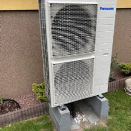 Zewnętrzna jednostka klimatyzacyjna Panasonic zamontowana na betonowych bloczkach przy ścianie budynku, z widocznymi rurami odprowadzającymi skropliny i dekoracyjnym podsypaniem z białych kamyków.