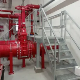 Metalowe schody z kratownicy prowadzące do platformy z czerwonymi rurami przemysłowymi i zaworami, widok na betonową podłogę i ścianę.