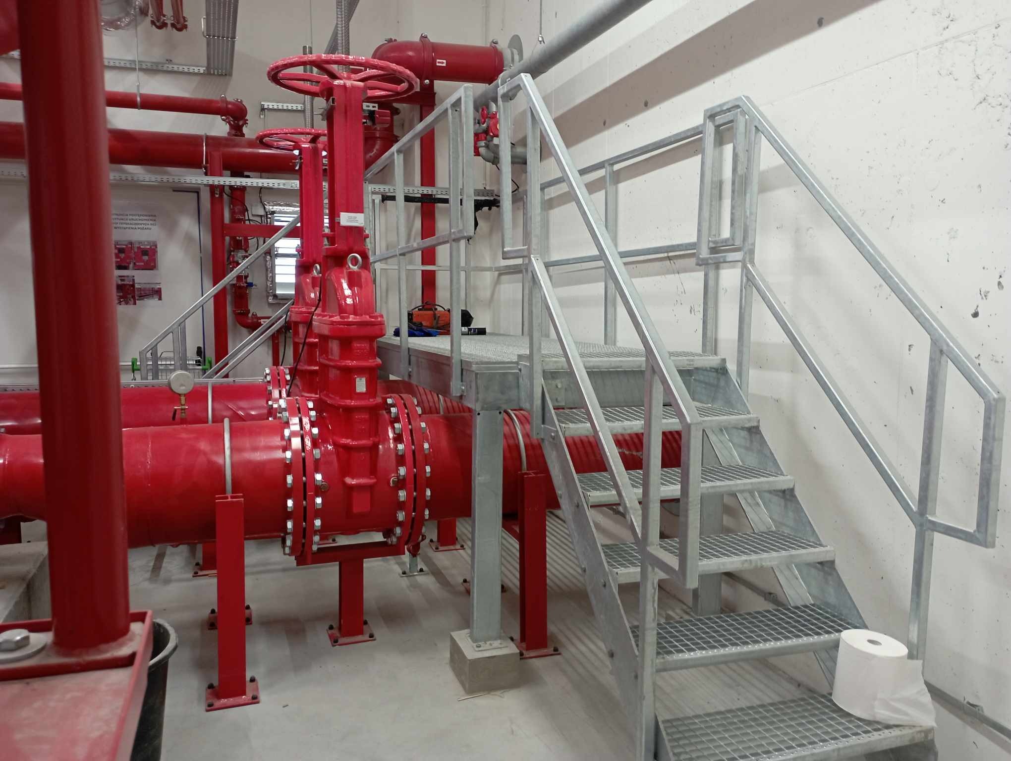 Metalowe schody z kratownicy prowadzące do platformy z czerwonymi rurami przemysłowymi i zaworami, widok na betonową podłogę i ścianę.