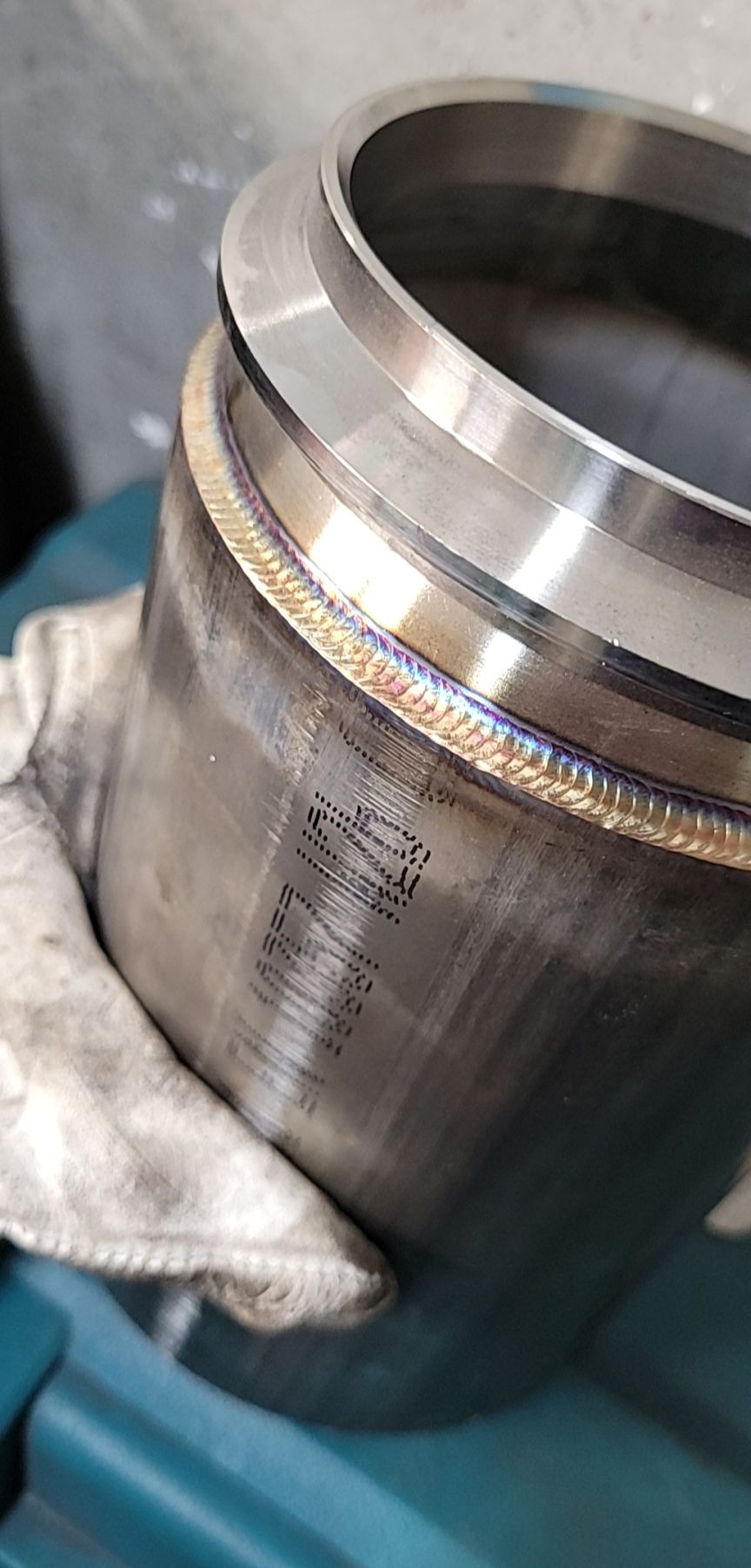 Metalowy cylinder z precyzyjnym spoinem, widoczne tęczowe zabarwienie ciepła, trzymany w rękawicy ochronnej, z naniesionym laserowo oznaczeniem.