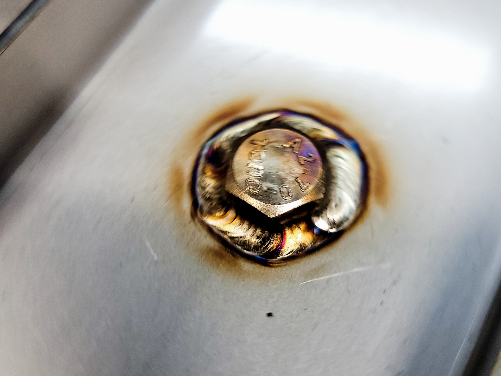 Zbliżenie spawu wokół śruby na metalowej powierzchni, widoczne ślady ciepła i charakterystyczne zabarwienia.