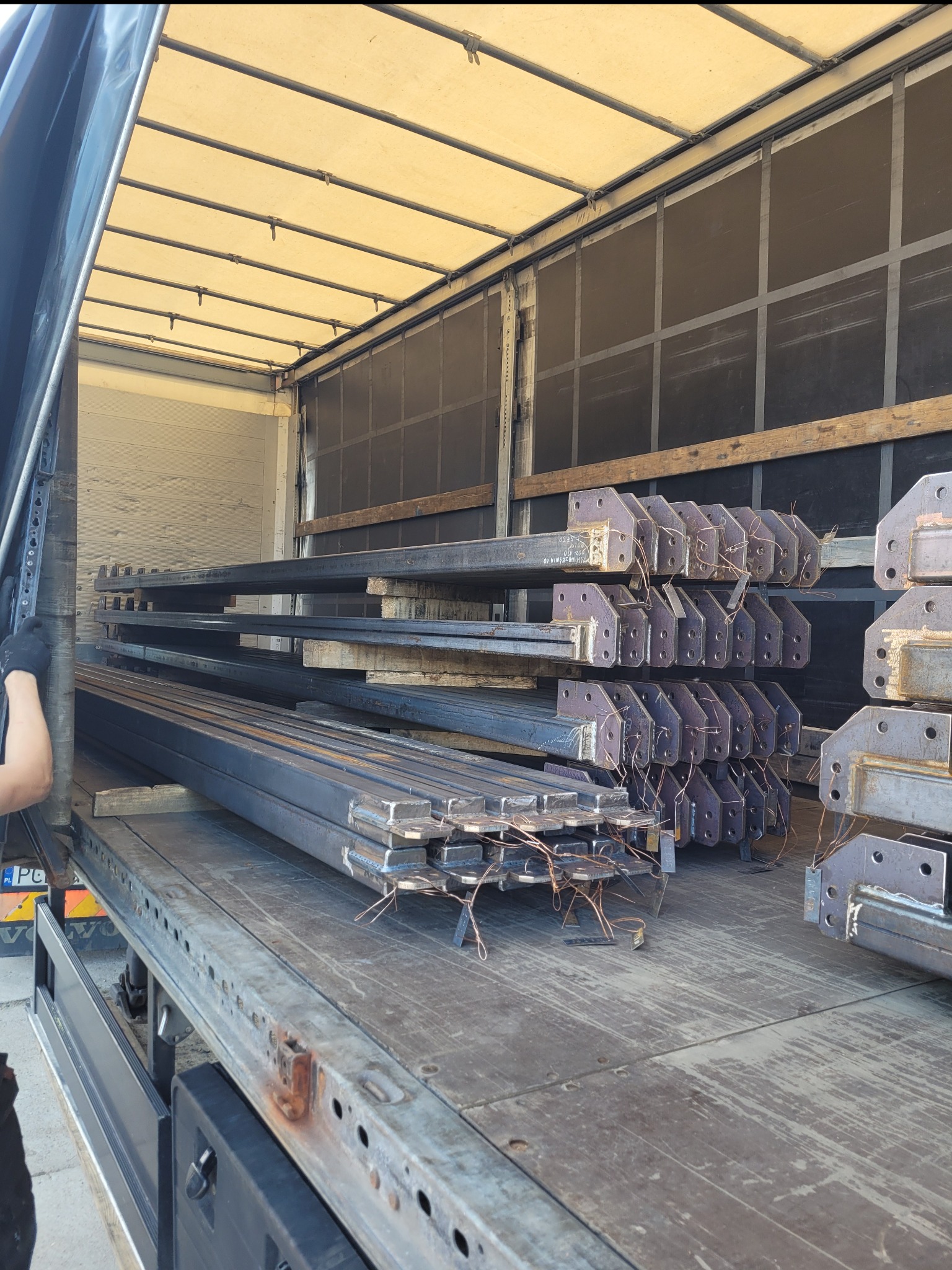 Transport elementów konstrukcji stalowej w naczepie, widoczne spawane profile z blachami mocującymi, zabezpieczone drutem spawalniczym.