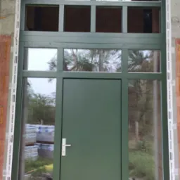 Nowoczesne, zielone drzwi zewnętrzne z przeszkleniami i nadświetlem w kształcie trapezu, obramowane taśmą montażową Soudal Window System, widoczne bloczki betonu kom&oacute;rkowego w tle.