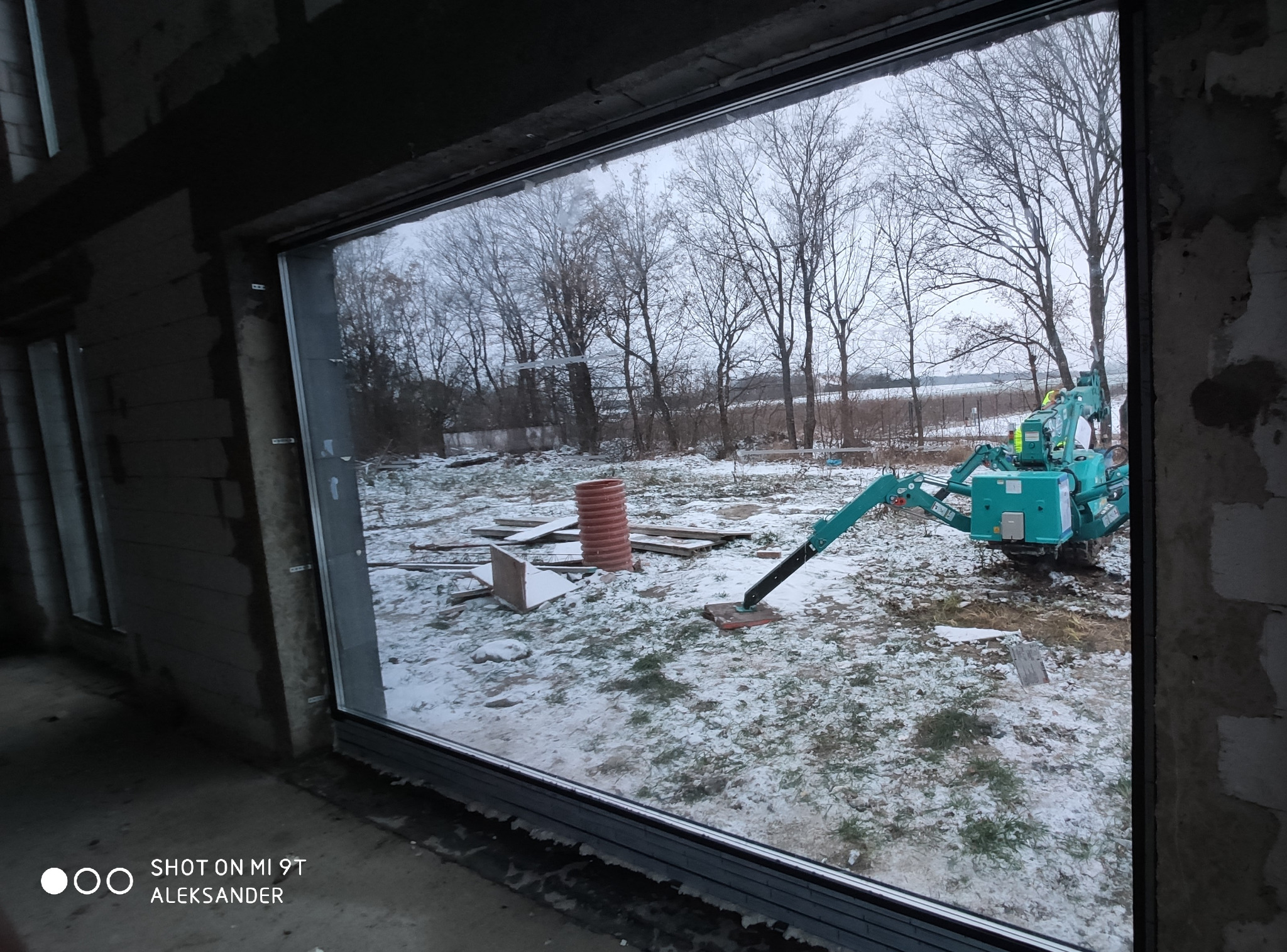 Widok z wnętrza budynku w stanie surowym na krajobraz zimowy przez nowo zamontowane okno PCV, na zewnątrz widoczna mała koparka i resztki materiałów budowlanych.