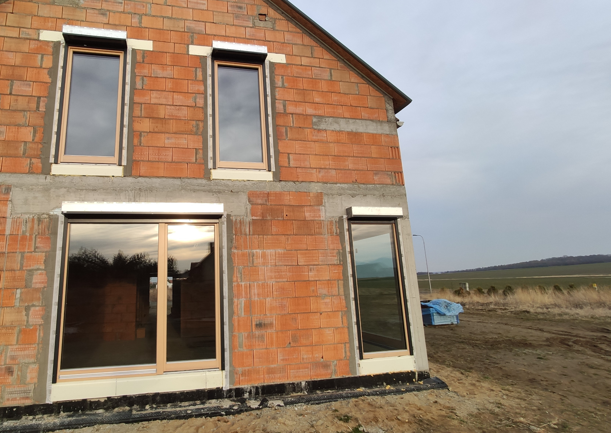 Surowy stan budynku z cegły z zamontowanymi oknami w beżowych ramach i roletami zewnętrznymi, widok na polną okolicę.