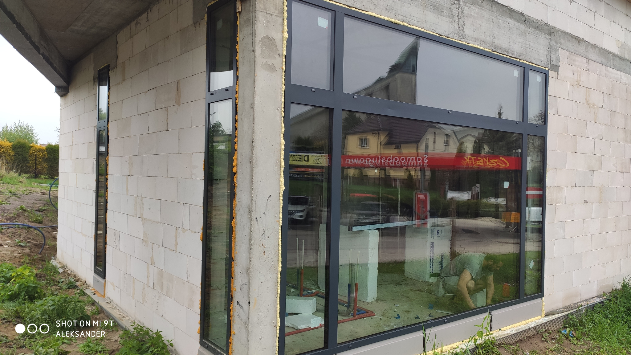 Montaż dużych okien w surowym budynku z betonu komórkowego, widoczna piana montażowa i odbicie otoczenia w szybach, pracownik wewnątrz budynku.