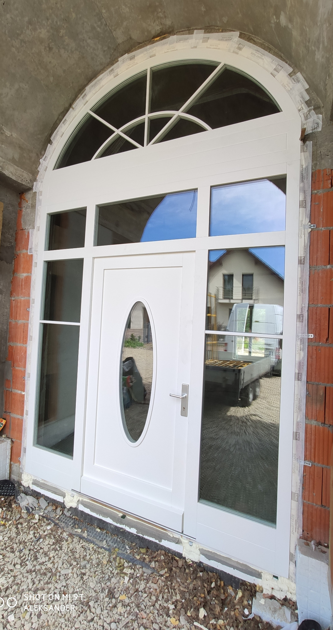 Białe drzwi wejściowe z owalnym przeszkleniem i łukowym nadświetlem, zamontowane w surowym stanie, widoczne ślady pianki montażowej i taśmy ochronnej.