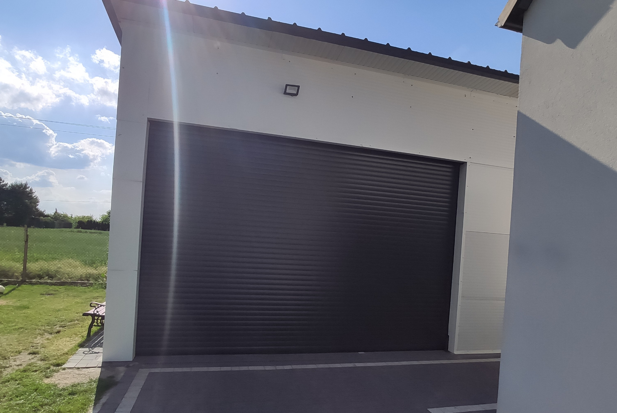 Zamknięta, grafitowa roleta zewnętrzna na tle białej ściany budynku z widocznym oświetleniem zewnętrznym i fragmentem zielonego ogrodu z ławką.