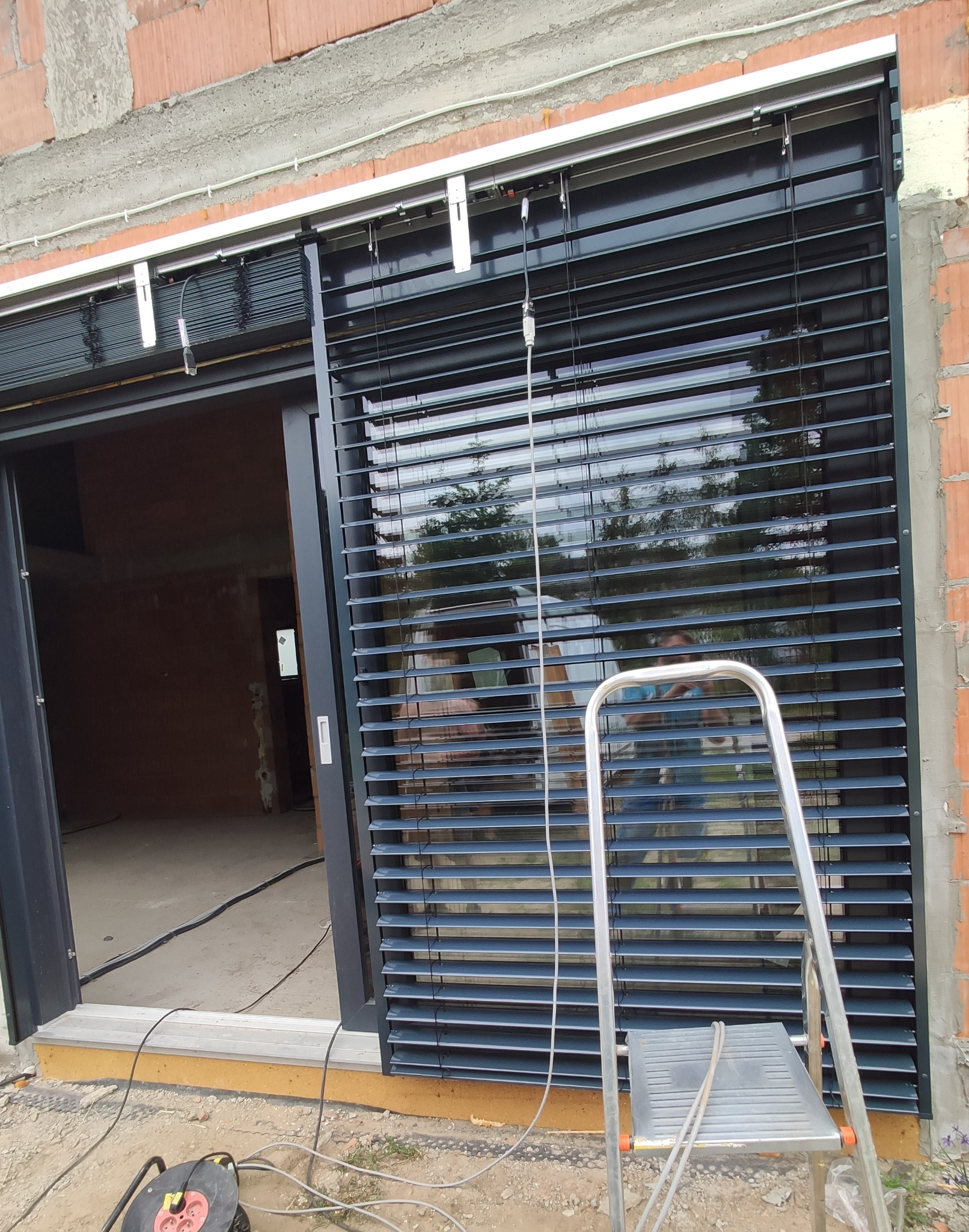 Montaż zewnętrznych żaluzji fasadowych w nowym budynku, widoczna drabina i przewody zasilające.