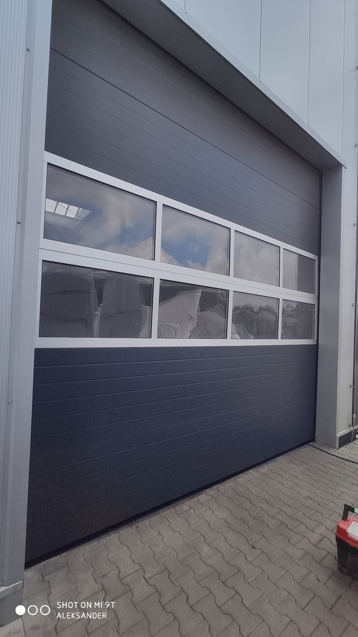 Szara segmentowa brama garażowa z rzędem okien z białymi ramami, widoczne odbicie nieba i zarys wnętrza budynku.