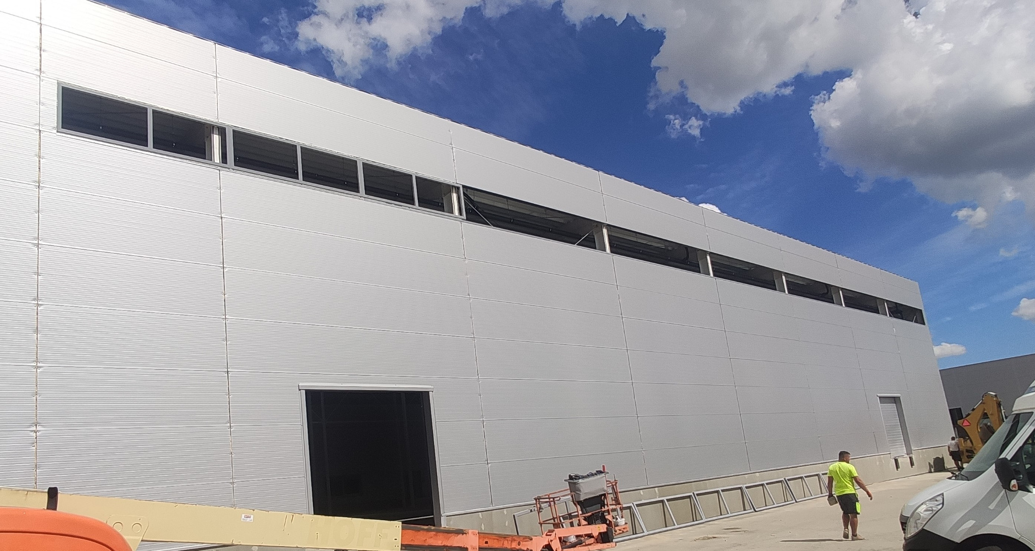 Montaż okien aluminiowych w hali przemysłowej z widoczną elewacją z paneli i częściowo zamontowanymi oknami w górnej części ściany, pod błękitnym niebem z chmurami.