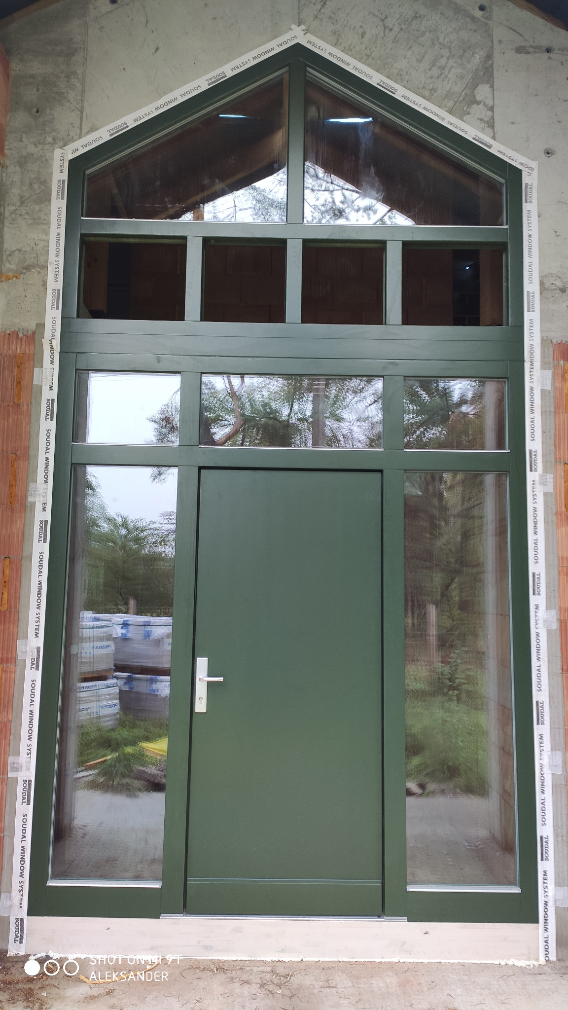 Nowoczesne, zielone drzwi zewnętrzne z przeszkleniami i nadświetlem w kształcie trapezu, obramowane taśmą montażową Soudal Window System, widoczne bloczki betonu komórkowego w tle.