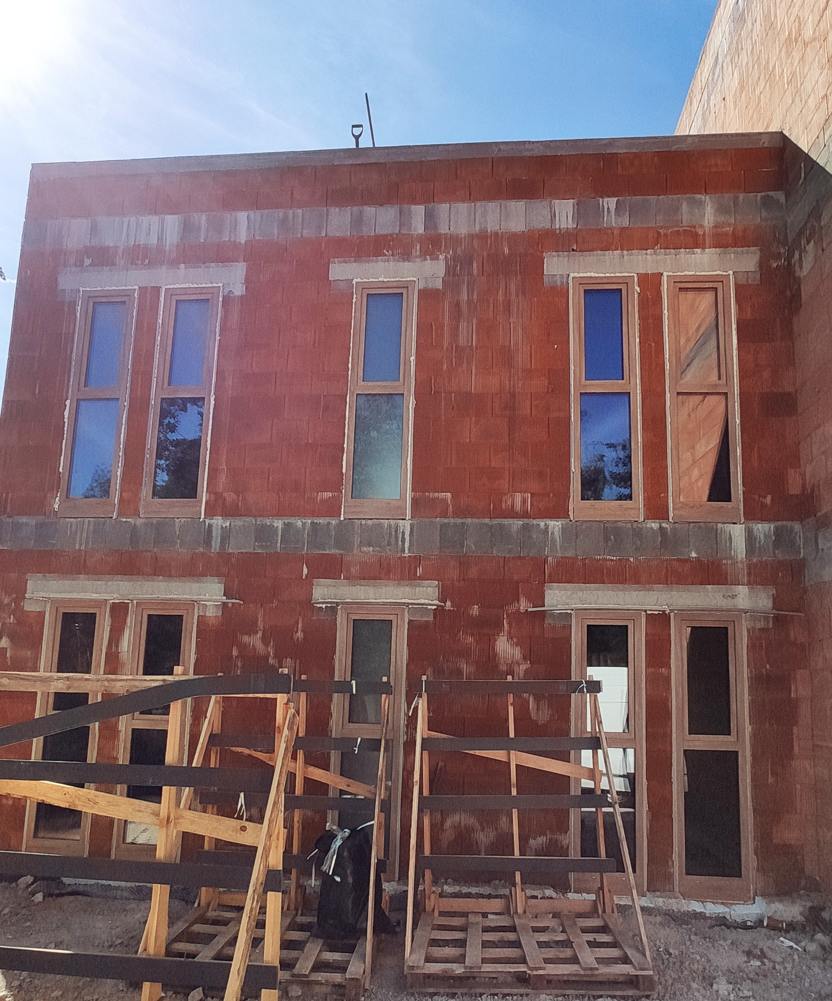 Surowy stan budynku z zamontowanymi oknami i drzwiami w drewnianych ramach, widoczne rusztowania i materiały budowlane.