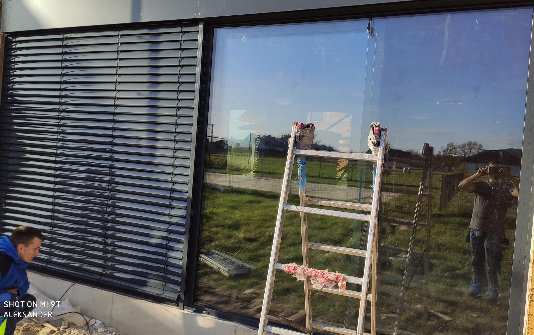 Montaż czarnych rolet zewnętrznych i okna, drabina oparta o szybę, widoczny monter oraz odbicie fotografa w szybie.