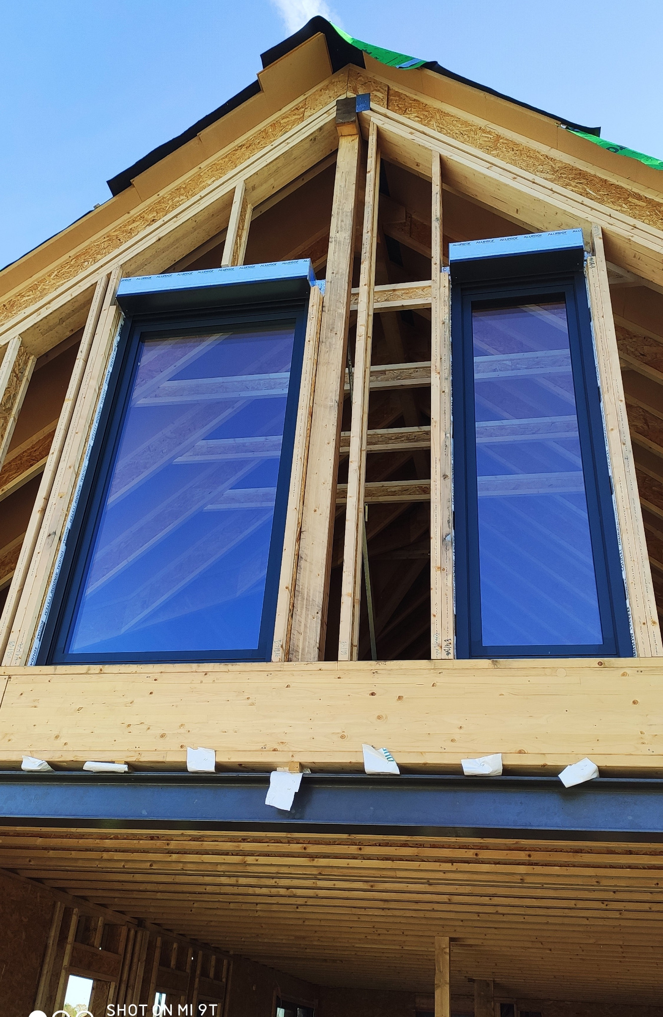 Surowy stan budowy domu z dwoma zamontowanymi oknami z roletami zewnętrznymi w kolorze antracytowym, widoczne elementy konstrukcyjne dachu i okien, niebieskie niebo w odbiciu szyb.