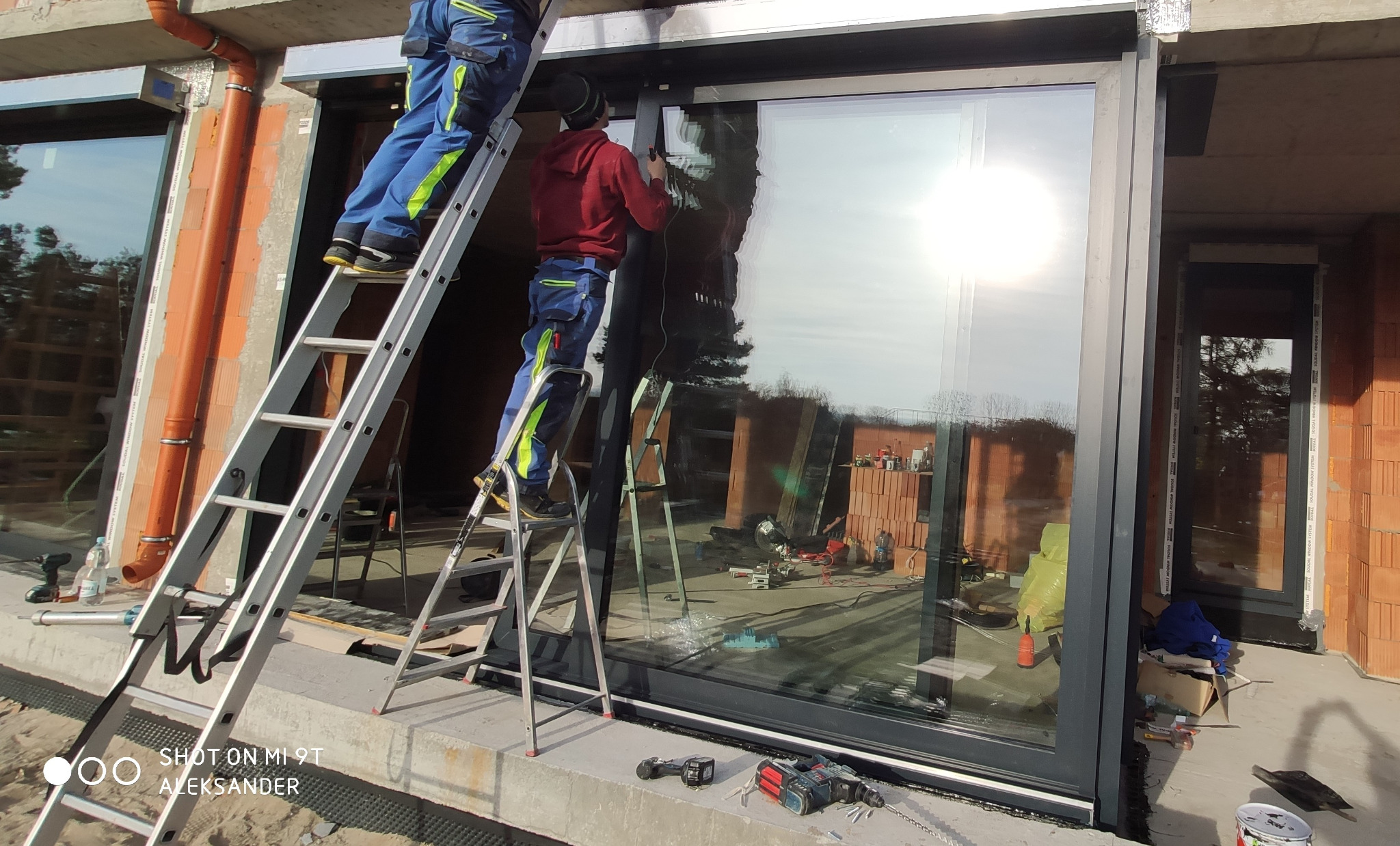 Montaż okna panoramicznego z czarną ramą przez dwóch pracowników na drabinach, widoczne narzędzia i surowy stan budynku.