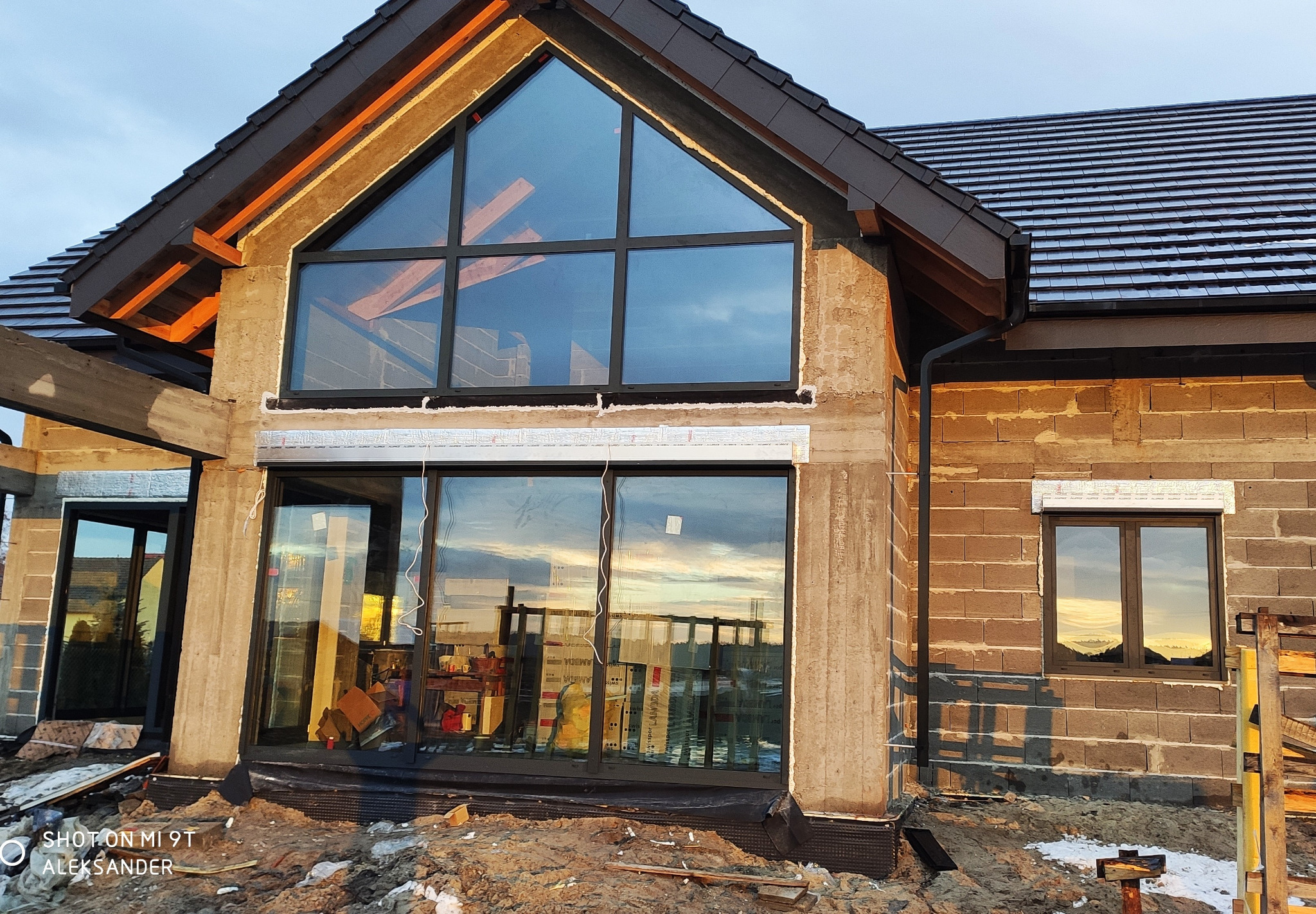Dom w trakcie budowy z zamontowanymi oknami z czarnymi ramami, widoczne okno trójkątne na poddaszu i duże okno przesuwne na parterze, w szybach odbija się zachodzące słońce.