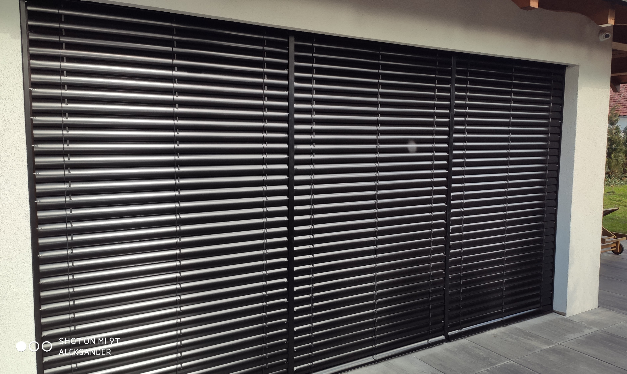 Zewnętrzne, poziome rolety w kolorze ciemnego grafitu, zamontowane na elewacji budynku z widocznym fragmentem kostki brukowej i zieleni w tle.