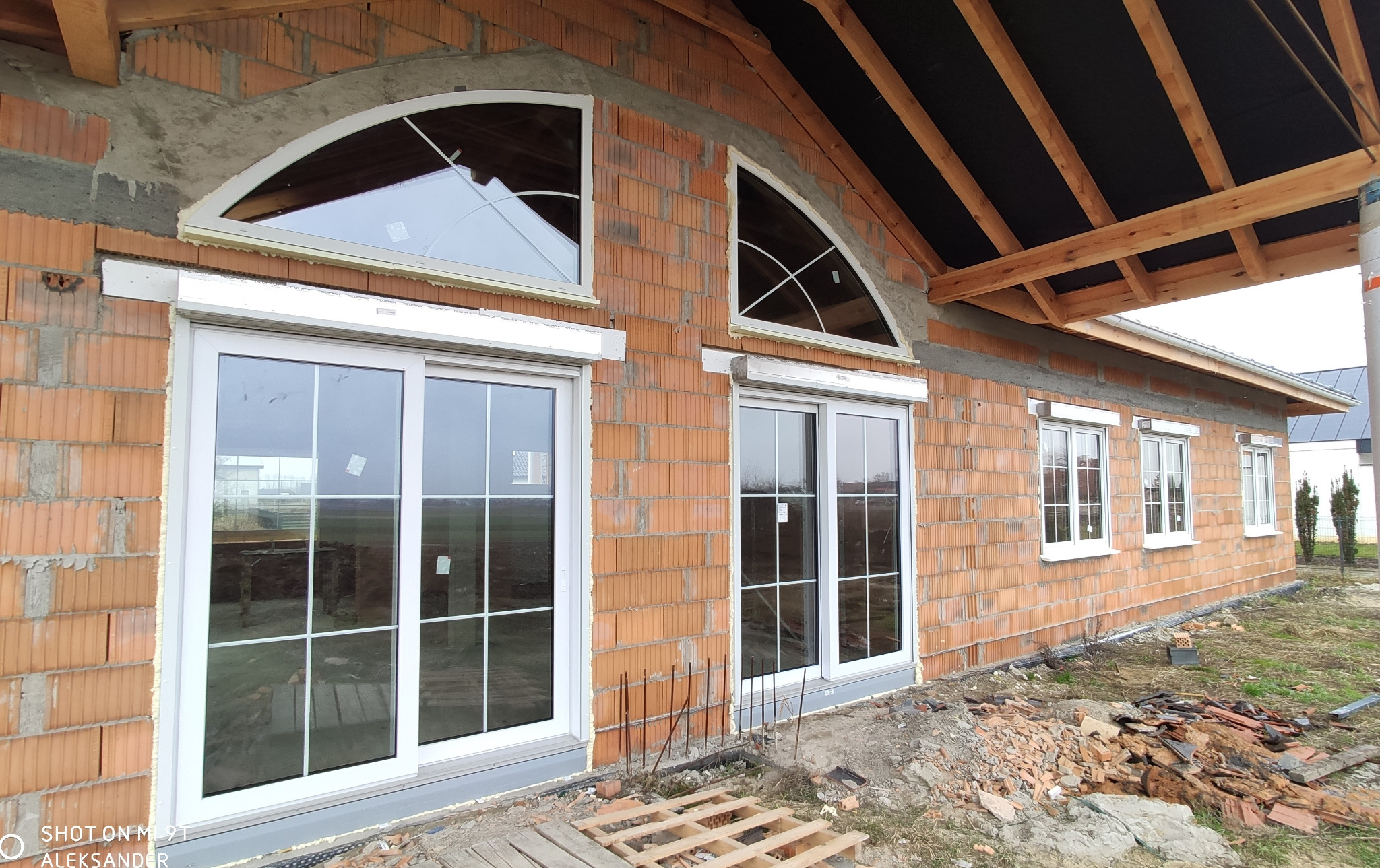 Surowy stan budynku z czerwonej cegły z zamontowanymi białymi oknami i roletami zewnętrznymi; widoczne dwa okna w kształcie półkola na górze.